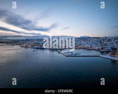 Aerial view of the city of Tromso covered in snow during winter, with the harbor, boats, and snow-covered buildings illuminated by soft twilight light Stock Photo