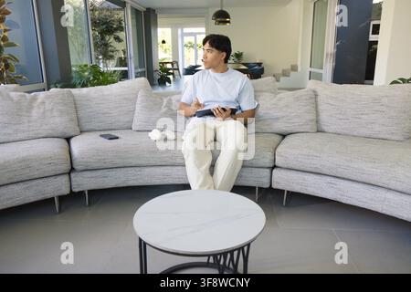 Asian man sitting on curved sofa using tablet with stylus, headphones and smartphone in living room. Minimalist, contemporary, modern, openplan, dayli Stock Photo