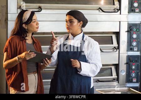 Female kitchen coworkers discussing baking, chef in apron pointing at ovens, manager using tablet. Bakery, collaboration, teamwork, professional, stai Stock Photo
