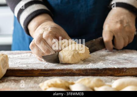 Hands of an elderly person portion soft dumpling dough using a sharp knife on a flour dusted surface, capturing the skill and warmth involved in creat Stock Photo