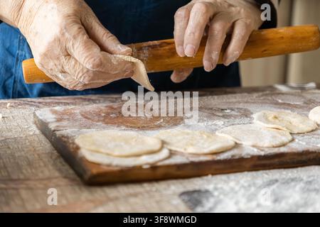 Elderly hands press dough with a wooden pin while making homemade dumplings on a floury cutting board, highlighting authentic cooking techniques and a Stock Photo