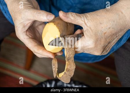 An elderly individual manually removes potato skin with a paring blade to prepare dinner, representing healthy homemade food production, slow living, Stock Photo