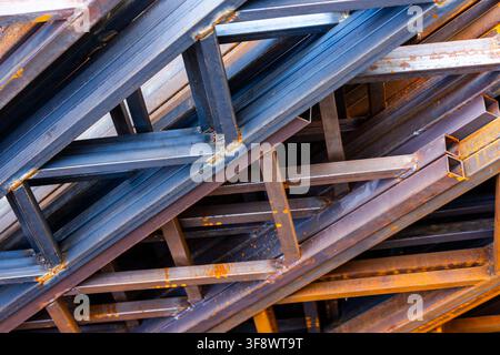 Close-up view of a chaotic pile of stacked metal beams. Dark blue-grey beams with weld marks contrast with oxidized rusty orange-brown pieces, showcas Stock Photo