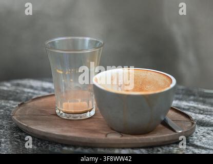 a finished matte-grey ceramic espresso cup with delicate crema residue alongside a clear water glass, elegantly presented on a rustic wooden saucer at Stock Photo