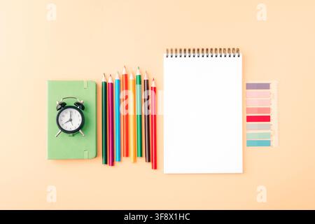 Colorful stationery with pencils, notebook, ruler and alarm clock arranged on a beige background with copy space. Top view, flat lay. Stock Photo