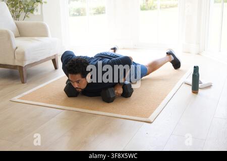 African American man doing push-ups on tan woven rug in living room, gripping black hex dumbbells Stock Photo