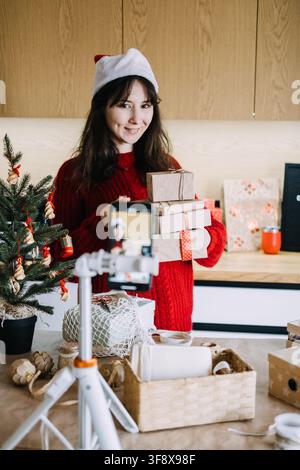 Smiling woman in Santa hat holds stack of wrapped parcels before phone on tripod in kitchen. Small business packing orders, handmade gift packaging Stock Photo