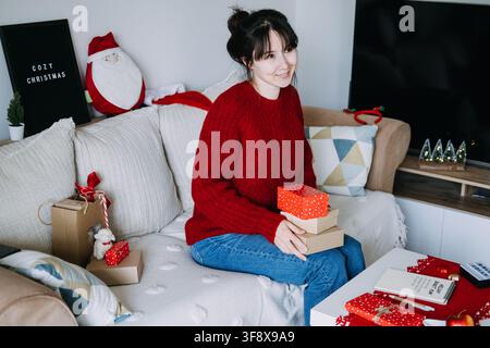 Smiling woman holds stack of wrapped boxes on sofa beside notebook with HOLIDAY BUDGET PLAN and COZY CHRISTMAS sign. Holiday budget planning notebook Stock Photo