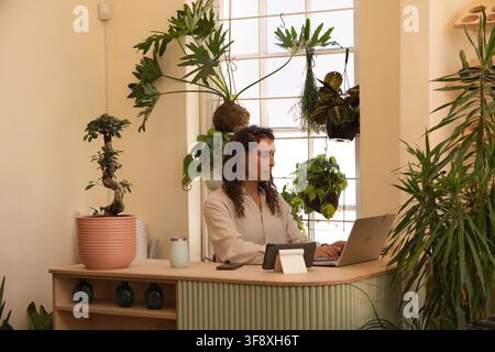 Nonbinary African American sitting at plant-filled curved counter using laptop, tablet, bonsai Stock Photo