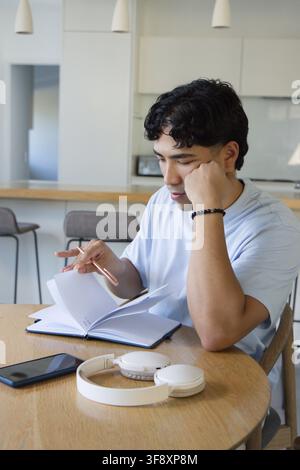 Asian adult man sitting at round table in home kitchen reading notebook, holding pen, headphones Stock Photo