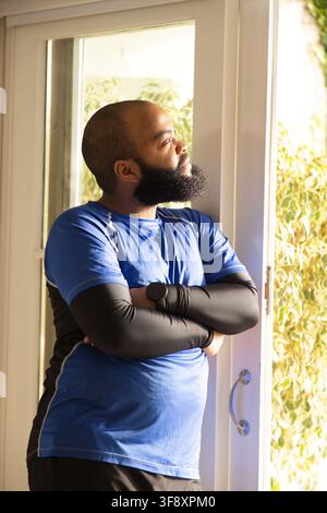 African American man standing arms crossed in blue shirt, smartwatch at home glass door looking out. Contemplative, sunlit, athleticwear, residential, Stock Photo