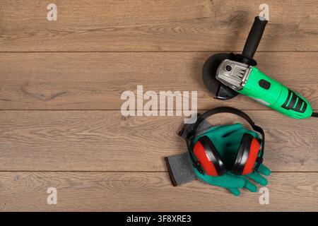 Top view of an electric angle grinder, protective work gloves and red earmuffs arranged on a rustic wooden surface. Industrial workplace safety equipm Stock Photo
