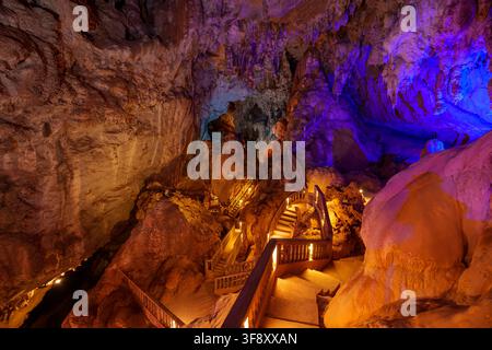Colored lights illuminate rock formations, stairways and walkways inside a large cave chamber. Purple, blue and orange lighting reveals the textured c Stock Photo