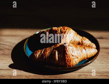 Two golden flaky croissants served on a dark ceramic plate on a wooden table, photographed in warm natural light Stock Photo