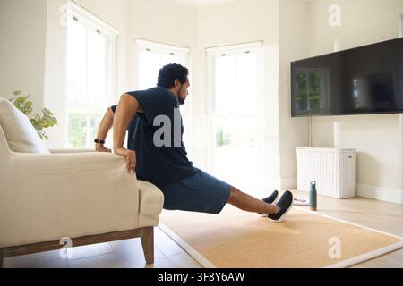 Mid-adult African American man doing tricep dips on beige sofa arm in living room, wrist tracker. Homefitness, workout, strength, minimalist, daylight Stock Photo