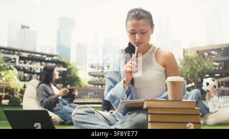 Focused student writing in notebook beside open book, taking notes and studying alone on campus lawn. Young woman does study session with stacked books and backpack. Stock Photo