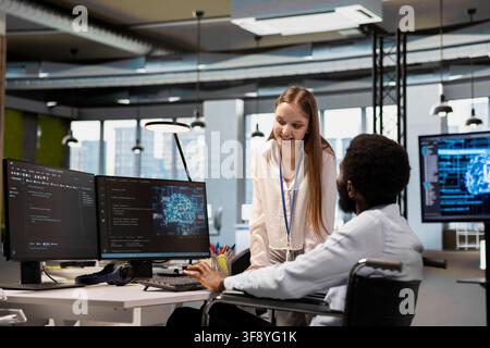 Cheerful senior programmer in wheelchair and junior dev using AI automation, refining algorithms. Joyful IT expert with paraplegia and young employee using artificial intelligence Stock Photo