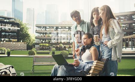 International students gather around laptop on campus bench, reviewing coursework notes and ideas together during outdoor learning collaboration. Group study project discussion. Stock Photo