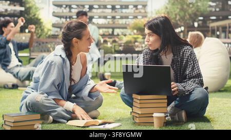 Diverse classmates collaborating on laptop while seated on campus lawn, discussing coursework and taking notes for exam preparation. Peer tutoring and group study. Stock Photo