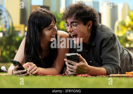 Young woman smiling at partner as they laugh at funny video content on smartphone, relaxing together on picnic blanket in park. Outdoor date with fun moment. Stock Photo