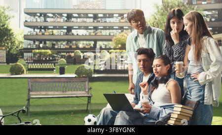 International students gather around laptop on campus bench, reviewing coursework notes and ideas together during outdoor learning collaboration. Group study project discussion. Stock Photo
