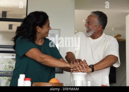 Senior African American couple laughing and pressing hands on white juicer at kitchen counter Stock Photo