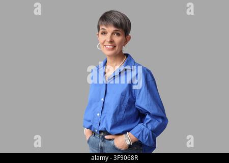 Smiling mature woman with short gray hair wearing a blue shirt and jeans, hands in pockets, on a gray background Stock Photo
