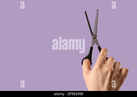 Hand holding open thinning shears for hair cutting and styling on a solid purple background, the metal scissors with black handles are positioned agai Stock Photo