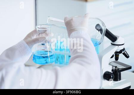 Scientist in white lab coat and gloves pouring blue liquid from a beaker into an erlenmeyer flask in a laboratory, Scientific research, chemical exper Stock Photo