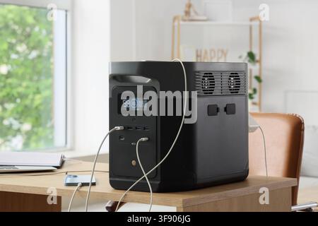 Portable power station charging multiple devices on a wooden desk in a modern home office setting, the modern black and gray unit features a digital d Stock Photo