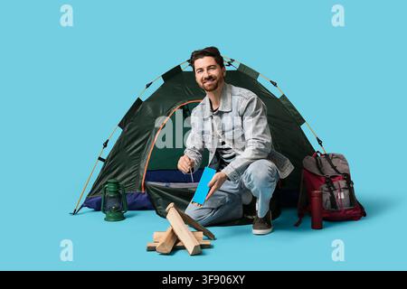 Young male tourist with tent and travelling equipment making fire on blue background Stock Photo