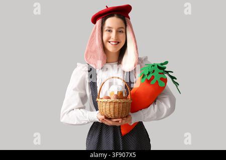 Stylish woman with bunny ears holding carrot toy and basket with Easter bread on grey background. Easter celebration Stock Photo