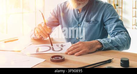 Senior man with white beard practicing calligraphy and brush lettering on paper at a wooden desk in a sunlit room, He sits at a wooden desk in a brigh Stock Photo