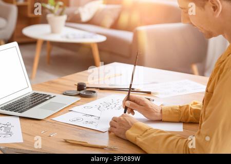 Young man practicing calligraphy and lettering at a wooden desk with a laptop in a sunlit home office, He uses a brush pen on paper, surrounded by var Stock Photo