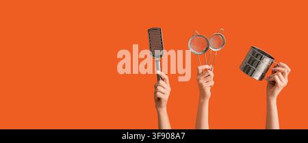 Hands holding kitchen utensils including a grater, two strainers, and a flour sifter on an orange background Stock Photo