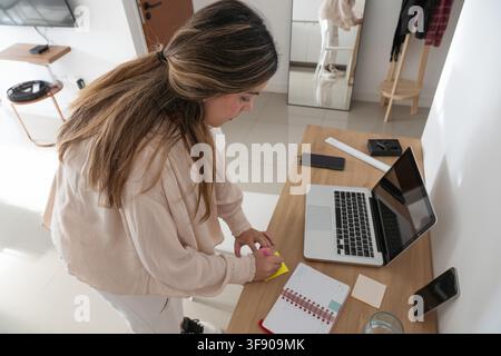 High angle overhead shot of woman writing on yellow sticky note at wooden desk, laptop open, mirror and coat rack visible in background Stock Photo