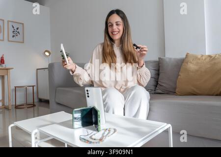 Cheerful woman sitting on couch holding up smartphone and lavalier microphone, phone with beaded strap propped on coffee table nearby Stock Photo