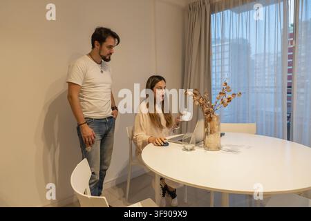 Woman sitting at table talking and gesturing near open laptop, man standing beside her listening, dried eucalyptus and window curtains in background Stock Photo