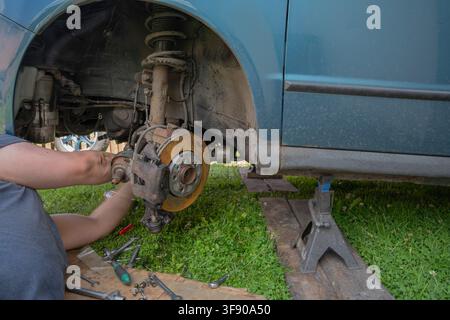 Mechanic working on a jacked-up teal car suspension system outdoors during DIY repair Stock Photo