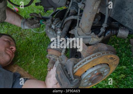 Mechanic lying on the ground using tools to repair car suspension and steering components outdoors Stock Photo