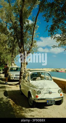 A vintage white Fiat 500 parked under a tree beside the water, Malta Stock Photo