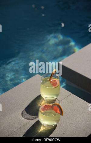 Two stemless glasses sitting on light-gray pool steps, green drink, citrus on rims, water shining. Glassware, grapefruit, outdoor, sunlight, reflectio Stock Photo