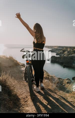 Bicycle woman coast Cyclist stands on a scenic cliff edge overlooking the ocean with her hand raised in a gesture of freedom Stock Photo