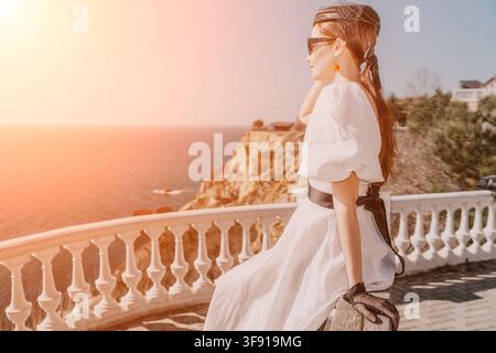 Woman white dress sunset sits on elegant stone balcony overlooking scenic Mediterranean coastline with copy space for luxury travel brands Stock Photo