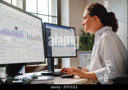 Focused professional businesswoman working with modern data analytics dashboards on multiple monitors in a sunlit office. The image conveys productivi Stock Photo