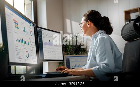 Professional businesswoman analyzing data charts, graphs, and performance metrics on dual monitors in a bright, modern office. The image captures a pr Stock Photo