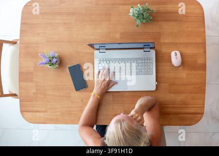 From above a man sits at a wooden table looking at a laptop screen while thinking. Remote work decision, home office freelance, thoughtful worker concept. Stock Photo