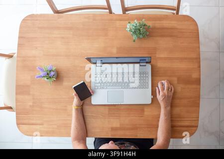 An overhead view of a man sitting at a wooden table with a laptop and holding a smartphone. Remote work lifestyle, home office freelance, modern workspace. Stock Photo
