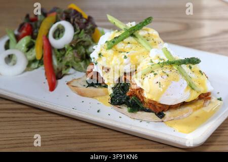 Eggs Benedict with pulled pork and wilted spinach on toasted English muffins, topped with hollandaise sauce and asparagus, served with a fresh side sa Stock Photo