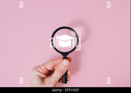 Hand holding magnifying glass over graduation hat icon on pink background, modern education research and academic career choice. High quality photo Stock Photo